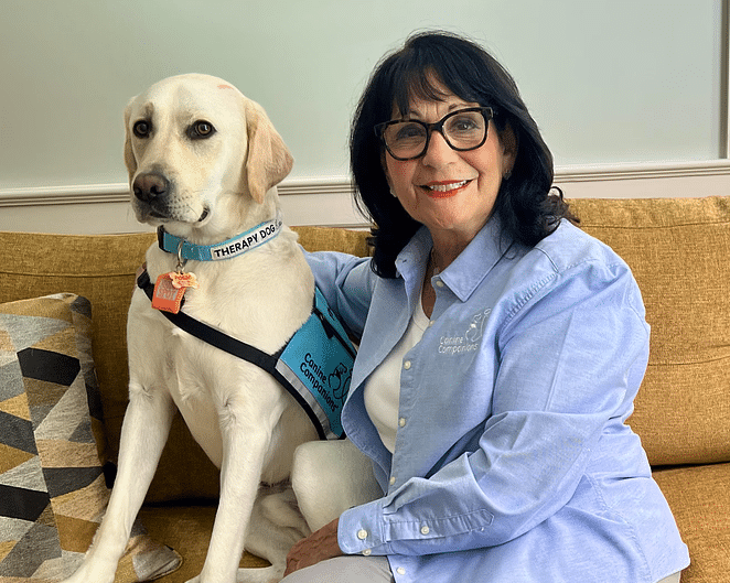 A woman, Yvonne, with glasses sits on a couch with a therapy dog, Picasso, in a blue vest beside her. 